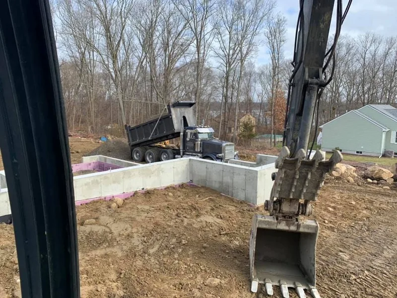 Excavator bucket beside a poured foundation wall with a dump truck in the background